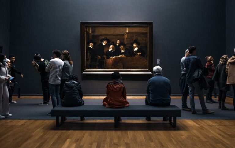Visitors sitting and standing in front of Rembrandt’s The Sampling Officials at the Rijksmuseum, in a dark gallery with deep blue walls.